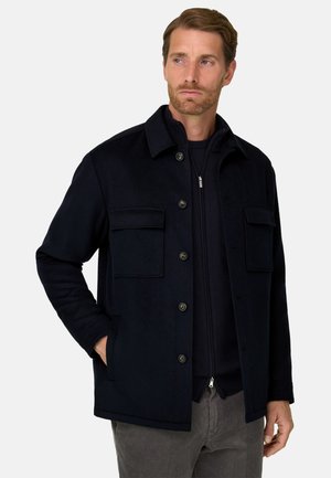 Un homme porte un manteau bleu marine foncé boutonné sur le devant, avec un col montant et des poches poitrine, associé à un pantalon gris sur un fond blanc uni.