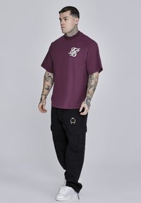 Camiseta de manga corta color marrón oscuro con logo, combinada con pantalones cargo negros que tienen bolsillos laterales y bordados en dorado. Zapatillas blancas completan el look.