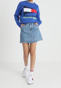 Modrá mikina s logem Tommy Jeans, džínová mini sukně, bílé tenisky s červenými detaily a pruhované ponožky. Sukně má roztrhaný lem.