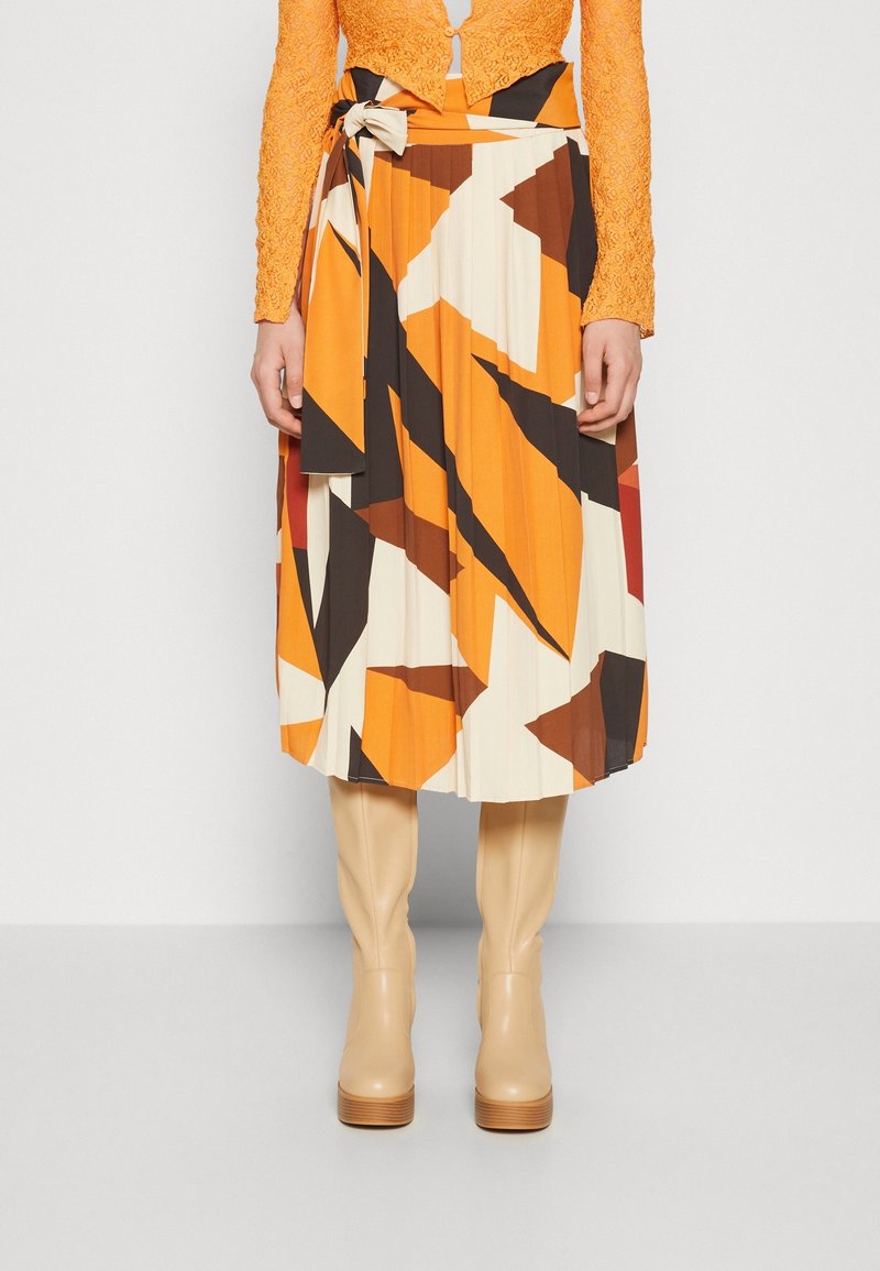 Jupe mi-longue plissée avec motif géométrique orange, marron, noir et crème, ceinture nouée à la taille, associée à des bottes beiges montantes et un top en dentelle orange.
