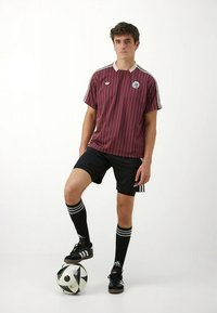 Vinröd randig fotbollströja med vit krage, matchad med svarta shorts och knälånga randiga strumpor. Spelaren står på en svartvitt fotbollsboll.