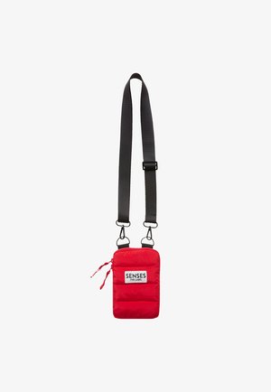 Red padded crossbody bag with adjustable gray strap, featuring a front pocket with a white label that reads "SENSES .THE LABEL."