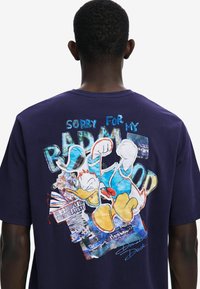 Fialové bavlněné tričko s grafickým potiskem Kačera Donalda a textem "Omlouvám se za svou špatnou náladu" na zádech. Design obsahuje barevné akcenty a textury.