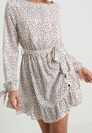 Robe blanche à manches longues avec petits pois noirs, ceinture nouée à la taille, ourlet à volants boutonné et nœuds aux manches, portée par une personne debout.