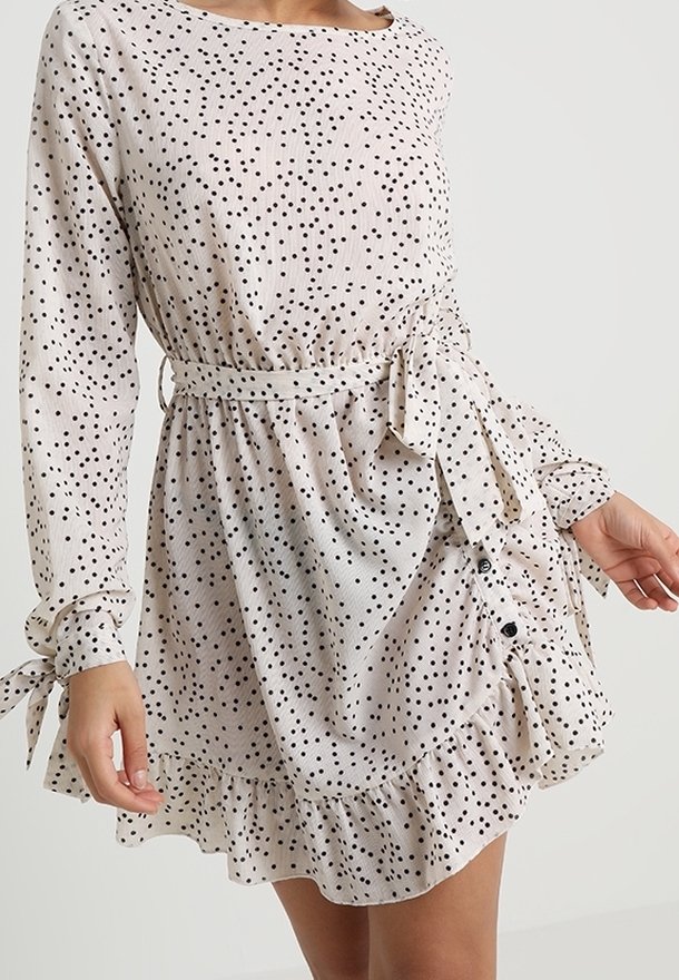 Robe blanche à manches longues avec petits pois noirs, ceinture nouée à la taille, ourlet à volants boutonné et nœuds aux manches, portée par une personne debout.