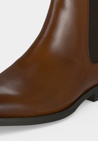 Bottine en cuir marron à finition lisse, panneau latéral élastique noir et semelle en caoutchouc noire.