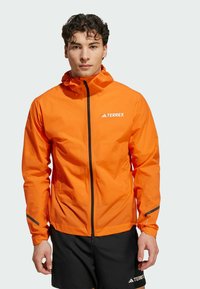 Veste imperméable orange avec capuche, fermeture éclair avant et accents réfléchissants. Fabriquée dans un matériau léger, elle présente un logo sur la poitrine.