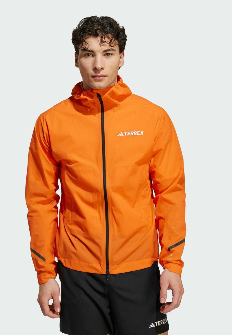 Veste imperméable orange avec capuche, fermeture éclair avant et accents réfléchissants. Fabriquée dans un matériau léger, elle présente un logo sur la poitrine.