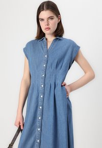 Abito in denim con maniche corte, chiusura a bottoni frontale e dettagli plissettati. Colore azzurro chiaro, texture liscia e silhouette rilassata.