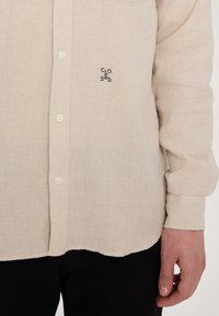 Helles beige Leinenhemd mit Knopfleiste und einem kleinen schwarzen "CLO"-Logo, das links aufgestickt ist. Lange Ärmel mit geknöpften Manschetten.