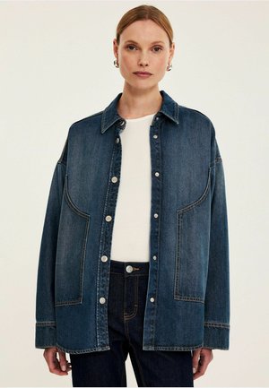 Kurtka dżinsowa w ciemnym niebieskim kolorze, z dużymi kieszeniami na naszywki, srebrnymi zatrzaskami i luźnym krojem z usztywnionym kołnierzem. Noszona na białej koszuli.
