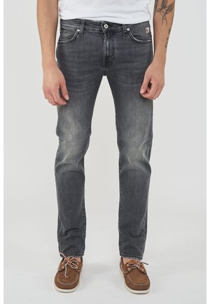 Uomo che indossa jeans neri sbiaditi, scarpe da barca marroni e una camicia bianca, con un tatuaggio visibile sul braccio sinistro.