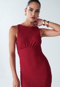 Punane tekstuuriga varrukateta kleit, millel on istuv vöökoht ja lehvikukujuline alumine serv. Aksessuaarideks on kuldsed kõrvarõngad ja käevõrud. Taust on lihtne.