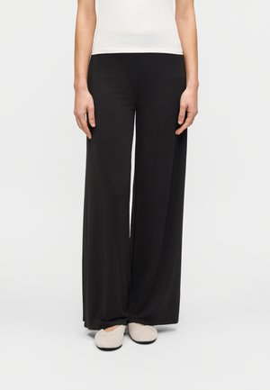 Fekete, bő szárú nadrág, puha, szellős anyagból, magas derékrész nélkül, világos színű bebújós cipőkkel párosítva.