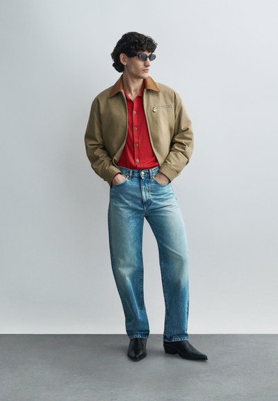 Braune Jacke mit einem beige Kragen, rotes Hemd mit Knöpfen, hellblaue Jeans und schwarze Stiefel. Das Model trägt dunkle Sonnenbrillen und steht vor einem neutralen Hintergrund.