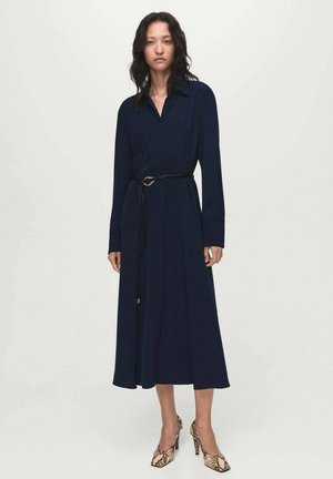 Woman wearing a long-sleeve navy midi dress with a thin black belt and snakeskin high heels, standing against a plain white background.