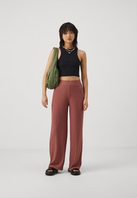 Schwarzes, kurzes Tanktop, gerippte, rostfarbene, weit geschnittene Hose, schwarze Plateausandalen und eine grüne, gesteppte Tasche. Geschichtete, bunte Perlenkette.