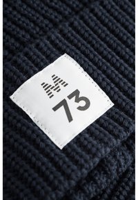 Tume sinine kootud kangas, millel on sooniline tekstuur, ning valge silt musta trükiga, millel on tähised "M" ja "73".