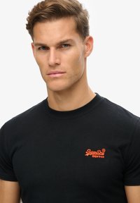 T-shirt en coton noir avec un col rond et des manches courtes, ornée d'un logo "SuperDry" rouge brodé sur la poitrine.