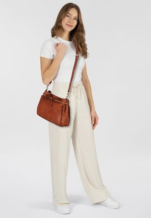 Borsa a mano in pelle marrone con dettaglio annodato e tracolla regolabile, mostrata con un modello che indossa una t-shirt bianca e pantaloni beige a gamba larga.