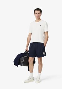 Camiseta de algodón blanca con un logo verde, pantalones cortos de malla azul marino con un gráfico, zapatillas blancas, llevando un bolso blanco y negro.