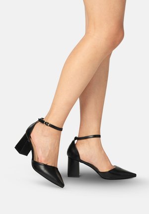 Legs wearing black pointed-toe block heels with ankle straps, posed on a neutral background.