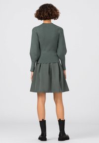 Zielony prążkowany sweter z bufiastymi rękawami w połączeniu z rozkloszowaną zieloną spódnicą, uzupełniony czarnymi botkami do kostki, widziany od tyłu.