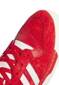 Zapatilla de ante roja con suela de goma blanca, que presenta rayas blancas, parte superior texturizada, cuello acolchado y detalles perforados en la puntera.