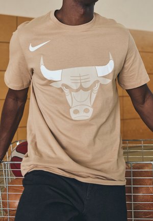 Mężczyzna w beżowej koszuli z białym logo byka i symbolem Nike stoi obok metalowego wózka na piłki do koszykówki.