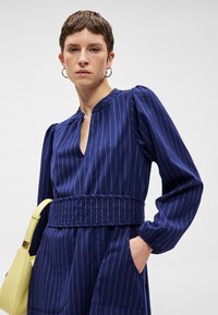 Tengerészkék ruha függőleges rózsaszín csíkokkal, V nyakkivágással, puffos ujjakkal és övvel a deréknál. Egy sárga kézitáska is látható.