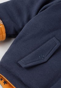 Dunkelblaue Stoffkleidung mit einer Klappentasche, die mit einem Druckknopf gesichert ist, und teilweise sichtbarem orangefarbenem Futter mit schwarzen Sternmustern.