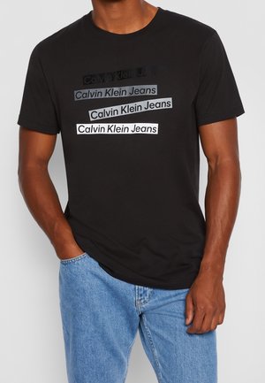 T-shirt nera in cotone con testo ripetuto "Calvin Klein Jeans" in diverse tonalità di grigio e bianco. Collo tondo classico e maniche corte.