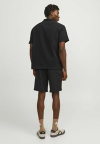 Ensemble homme composé d'une chemise noire à manches courtes et d'un short, en tissu texturé, coupe décontractée, accompagné de chaussettes beiges et de baskets, vu de l'arrière.