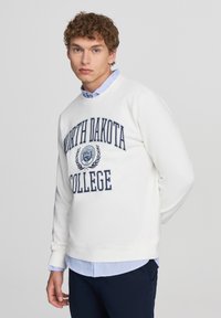 Felpa bianca con stampa blu navy "NORTH DAKOTA COLLEGE", dotata di colletto rotondo e polsini elasticizzati, indossata sopra una camicia collare azzurra chiara.