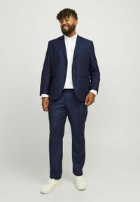 Navy suit made of smooth fabric, featuring a single-breasted jacket, two front pockets, and matching trousers. Paired with white sneakers.