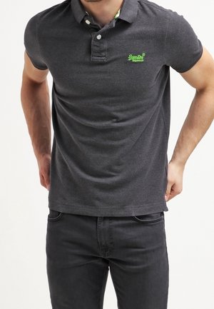 Mann in einem dunkelgrauen Poloshirt mit kleinem grünem Logo auf der linken Brust und schwarzen Jeans, steht mit den Händen in der Nähe der Hüften.