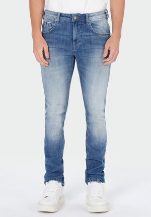 Man wearing slim-fit blue faded jeans and white sneakers, standing against plain background.