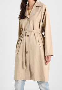 Donna che indossa un trench coat beige con cintura, chiusure a pulsante e tasche laterali, abbinato a jeans strappati di un blu chiaro.