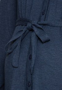 Vestido de tecido texturizado azul marinho com uma cintura cingida, apresentando botões e um decote com gola. Detalhe em close dos fechos.