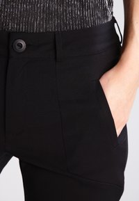 Pantalon noir avec une texture lisse, doté d'une fermeture à bouton, de poches latérales et d'une coupe ajustée. Le matériau semble flexible.