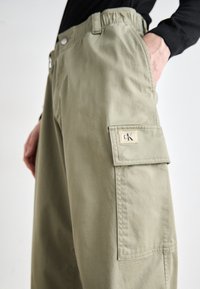 Pantalones cargo en tela verde oliva, con un bolsillo lateral con una etiqueta crema, cinturilla elástica y un diseño de corte relajado.