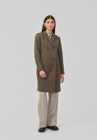 Dubbelzijdige bruine wollen jas met notch-lapels en zijzakken, gedragen over lichtgekleurde broeken en zwarte schoenen tegen een grijze achtergrond.