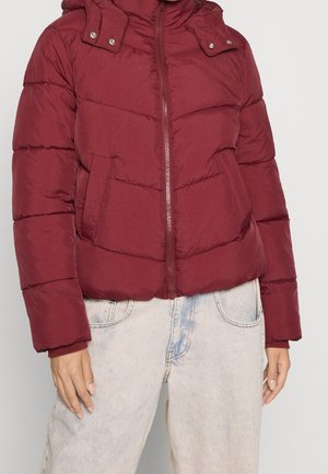 Osoba w bordowej pikowanej kurtce z przednimi kieszeniami i jasnych dżinsach, stojąca na tle jednolitego jasnego tła.