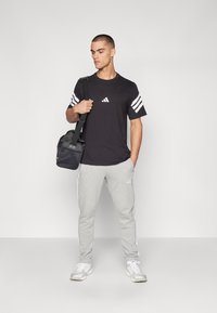 Svart t-skjorte med hvit tre-stripe-design på ermene, grå joggebukser med hvite sidestriper, og en svart treningsbag. Hvite joggesko.