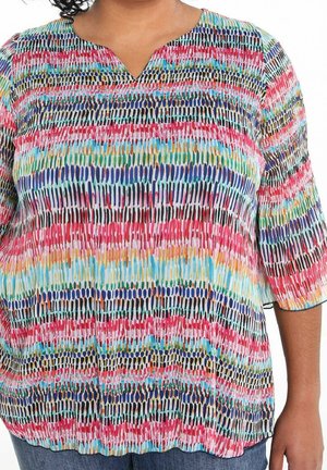 Vrouw draagt een meerkleurige gestreepte blouse met een kleine V-hals en driekwart mouwen, gecombineerd met een blauwe spijkerbroek.