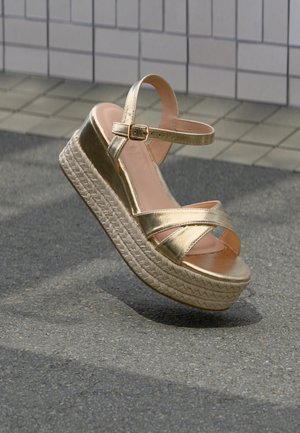 Zlatý platformový sandál s překříženými pásky, přezkou u kotníku a podešví z opleteného lana, umístěný ve vzduchu na asfaltu u obkladové stěny.