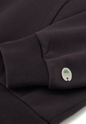 Sweat-shirt gris foncé en tissu doux et texturé. Présente des poignets côtelés et un petit logo brodé dans des couleurs neutres sur la manche.