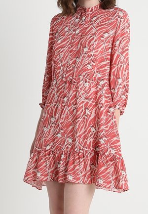 Kobieta ubrana w koralowo-różową sukienkę w kwiaty z długimi rękawami, zapięta na guziki z przodu, z paskiem do wiązania w talii i falbaną na dole.
