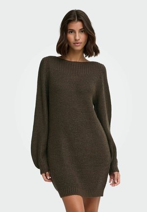 Femme aux cheveux courts et foncés portant une robe-pull en maille marron foncé à manches longues, debout devant un fond clair uni.