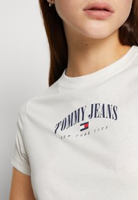 Bílý bavlněný tričko s námořnickým modrým nápisem "TOMMY JEANS" a malým logem. Kulatý výstřih, krátké rukávy, hladká textura.